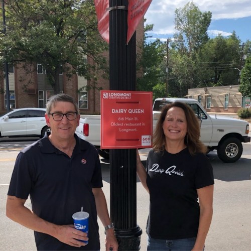 A man and woman stand by a sign for Dairy Queen, the oldest restaurant in Longmont, with a truck in the background.