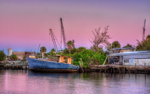 Abandoned boat on a calm waterway, surrounded by greenery and industrial cranes under a pastel sunset sky.