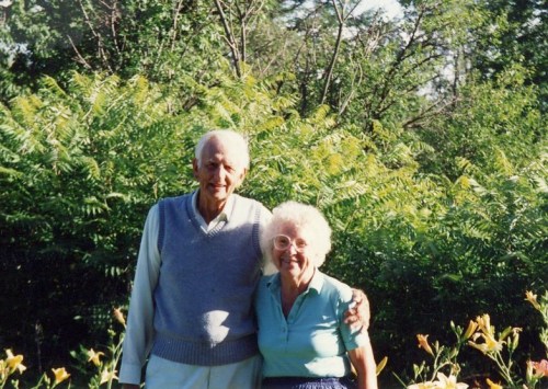 An elderly couple stands together outdoors, smiling, with lush greenery in the background.