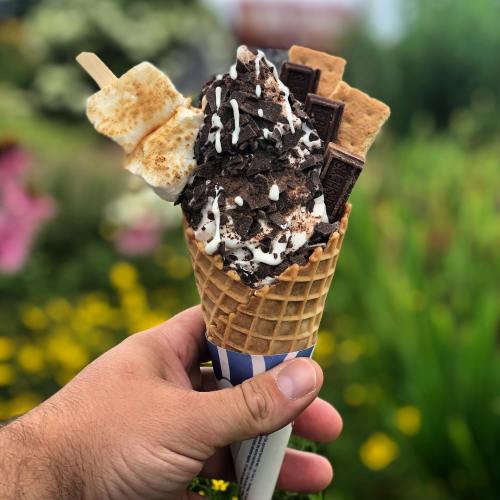 A hand holds a waffle cone filled with ice cream, topped with crushed cookies, chocolate bars, and a toasted marshmallow.