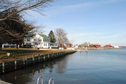 A serene waterfront scene featuring a white house, trees, and docks along a calm river under a clear blue sky.