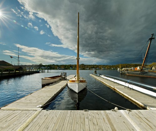 A calm marina scene with sailboats docked, under a dramatic sky with dark clouds and sunlight breaking through.