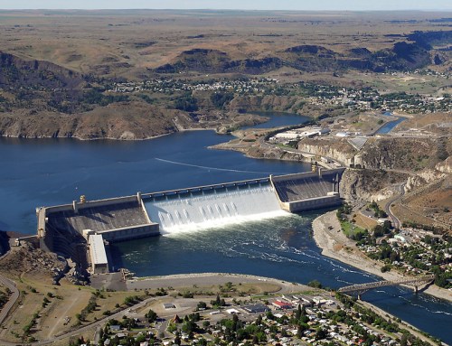 Aerial view of a large dam releasing water into a river, surrounded by hills and a small town in the distance.