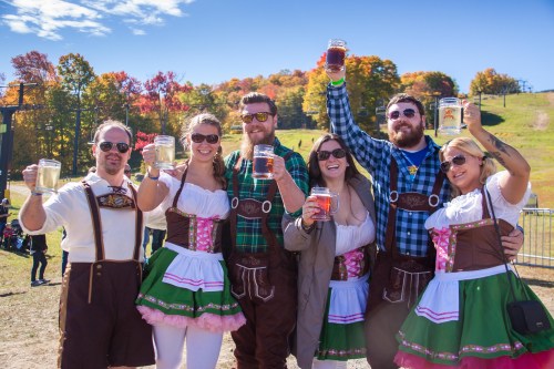 A group of six people in festive attire holding drinks, smiling outdoors with colorful autumn trees in the background.