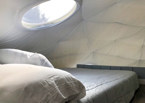 Cozy interior of a tent-like structure with a round window and a neatly made bed. Soft, light-colored bedding.