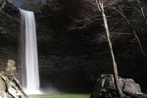 A serene waterfall cascades into a green pool, surrounded by rocky cliffs and bare trees.