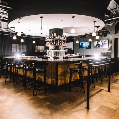 A modern bar with a circular counter, shelves of liquor, and pendant lights in a stylish, dimly lit setting.