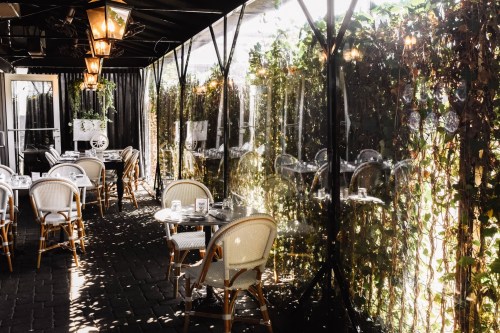 A cozy restaurant patio with tables, chairs, and lush greenery reflecting in the glass, illuminated by warm lanterns.