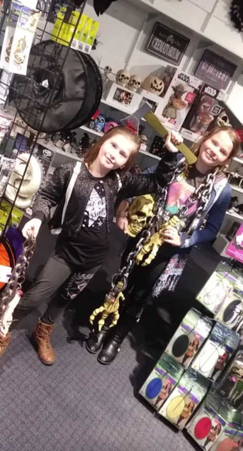 Two girls pose in a store filled with Halloween decorations, holding chains and a skull, surrounded by spooky items.