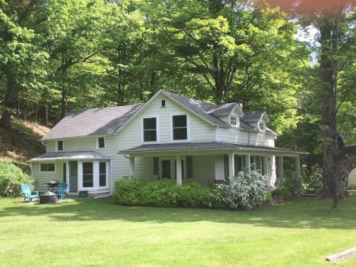 A white two-story house surrounded by lush green trees and a well-maintained lawn, featuring a porch and outdoor seating.