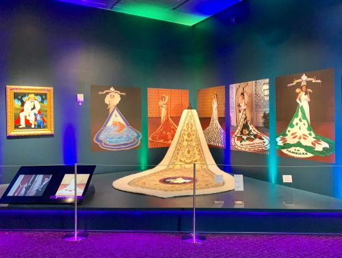 Art exhibit featuring colorful dresses and paintings, with a central display of a gown and illuminated walls.