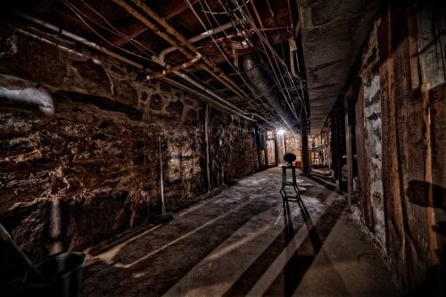 A dimly lit basement with stone walls, exposed pipes, and a single chair casting shadows on the floor.
