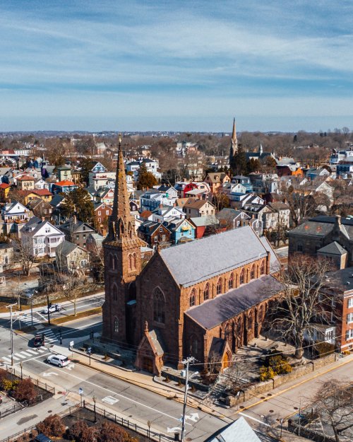 Aerial view of a historic church with a tall spire, surrounded by colorful houses and a clear blue sky.