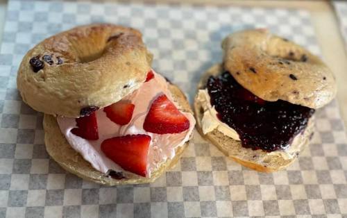 Two bagels filled with cream cheese, strawberries, and jam, served on a checkered paper background.