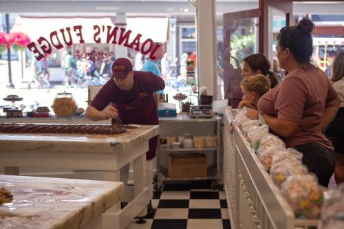 A candy maker shapes fudge while customers watch eagerly in a colorful candy shop.