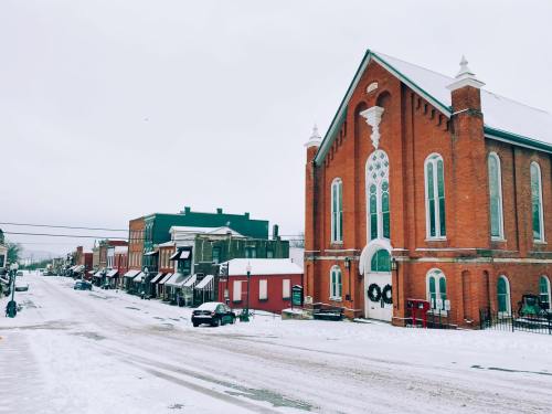 A snowy street scene featuring a brick church and quaint shops lined along the road in a small town.