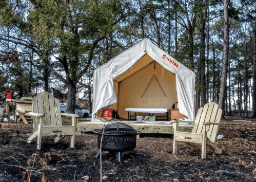 A camping tent set up in a wooded area, with two wooden chairs and a fire pit in front.