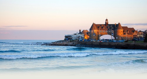 A coastal view featuring a historic building by the shore, with gentle waves and a pastel sky at sunset.