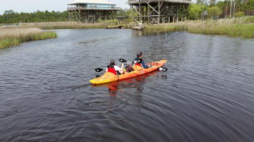 Two people kayak in a calm waterway surrounded by tall grass and wooden structures in the background.