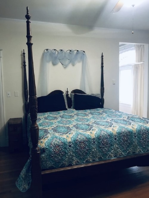 A four-poster bed with a patterned blue and green quilt, featuring a sheer canopy and two dark pillows.
