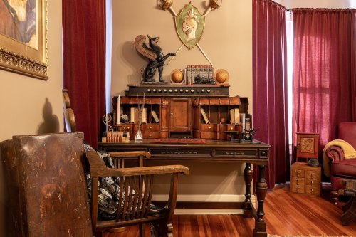 A vintage desk with decorative items, a wooden chair, and rich red curtains in a cozy room setting.