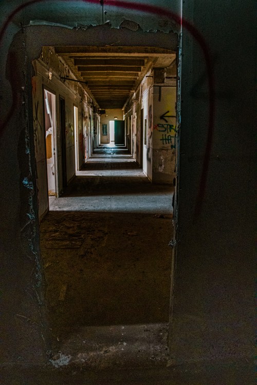 A dimly lit corridor with multiple doorways, featuring graffiti on the walls and a sense of abandonment.