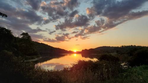 Sunset over a tranquil lake, surrounded by trees and hills, with colorful clouds reflecting on the water's surface.