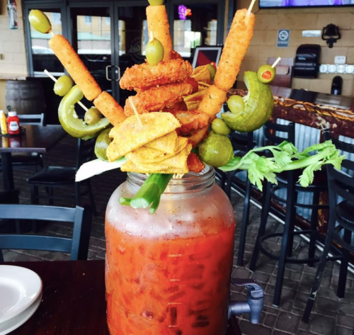 A large, colorful drink garnished with skewers of vegetables, olives, and fried snacks, served in a mason jar.