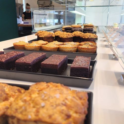 A display of assorted baked goods, including muffins and brownies, arranged on black trays in a bright bakery setting.