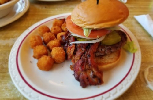 A burger with bacon, lettuce, tomato, pickles, and onions, served with a side of golden tater tots on a plate.