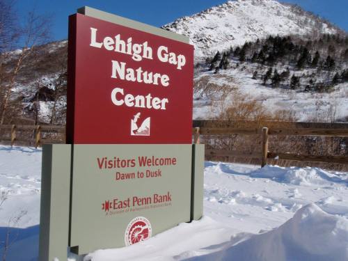 Sign for Lehigh Gap Nature Center, welcoming visitors from dawn to dusk, with snowy mountains in the background.