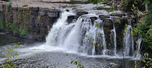 A serene waterfall cascading over rocky ledges into a calm river, surrounded by lush greenery.