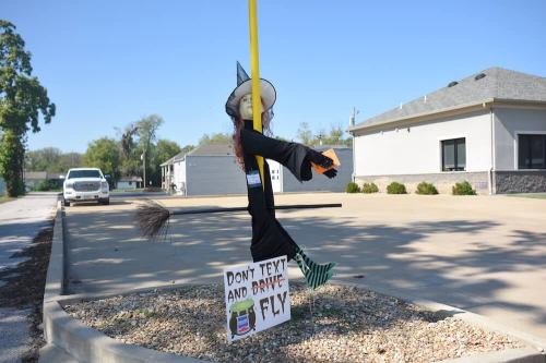 A scarecrow dressed as a witch hangs from a pole with a sign reading "Don't text and drive, fly."