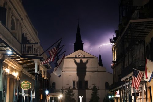 A nighttime scene of a street with historic buildings, flags, and a dramatic lightning strike illuminating a church steeple.