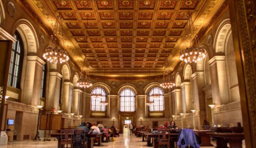 A grand library interior with ornate ceilings, chandeliers, and people studying at wooden tables.