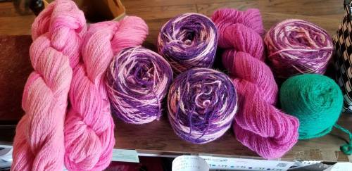 A collection of yarn in various shades of pink and purple, including skeins and rolled balls, on a wooden surface.