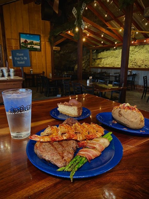 A table with a glass of water, a steak, bacon-wrapped shrimp, asparagus, a baked potato, and a slice of cheesecake.