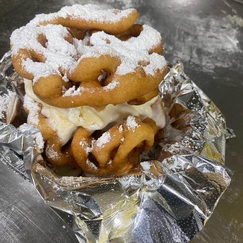A stack of funnel cakes layered with ice cream and dusted with powdered sugar, served on a foil plate.