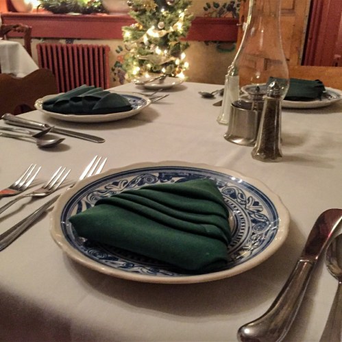 A dining table set with plates, green napkins, and silverware, decorated with soft lighting and festive greenery.