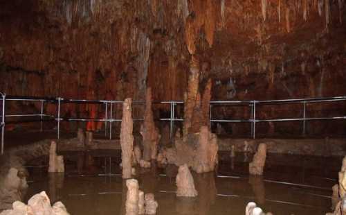 A dimly lit cave interior with stalactites and stalagmites, featuring a reflective pool of water and a safety railing.