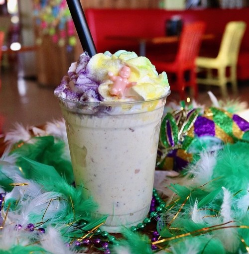 A colorful milkshake topped with whipped cream, a small figurine, and surrounded by green feathers and festive decorations.