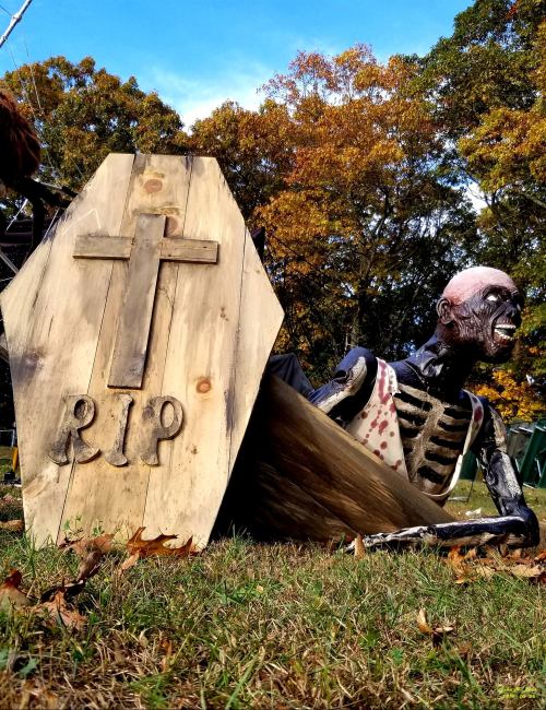 A skeleton emerging from a coffin with a wooden cross marked "RIP," set in a grassy area with trees in the background.