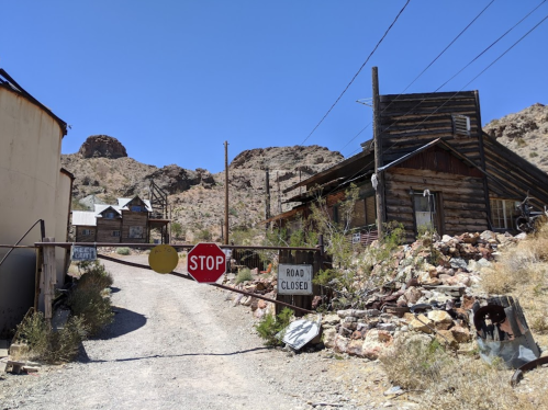 A deserted road in a ghost town, featuring a stop sign and closed road signs, with old wooden buildings and mountains in the background.