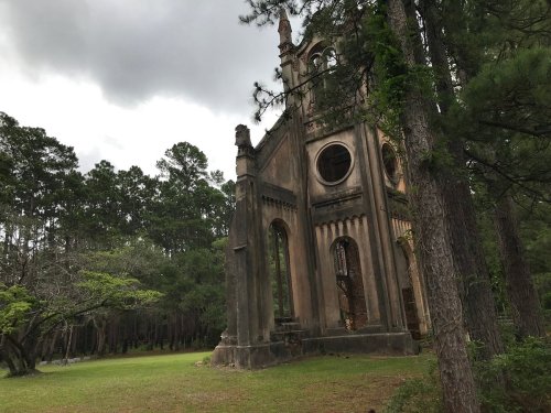 A partially collapsed stone structure surrounded by trees, set against a cloudy sky.