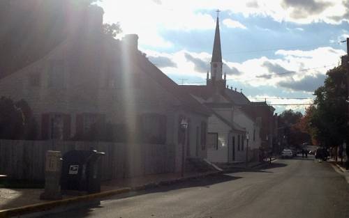 A quiet street lined with houses, featuring a church steeple in the background under a partly cloudy sky.