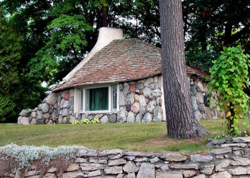 A quaint stone cottage with a unique roof, surrounded by trees and a stone wall in a lush green setting.