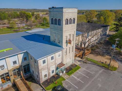 Aerial view of a historic building with a tall tower, surrounded by trees and a parking lot.
