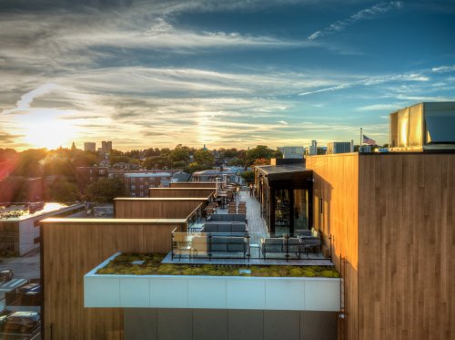 A rooftop terrace with seating overlooks a city skyline at sunset, featuring wooden architecture and lush greenery.