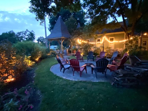 A cozy outdoor seating area with colorful chairs, surrounded by greenery and soft lights, at dusk.