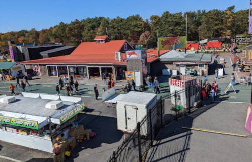 A vibrant outdoor fairground with food stalls, ticket booth, and visitors enjoying the attractions under a clear blue sky.